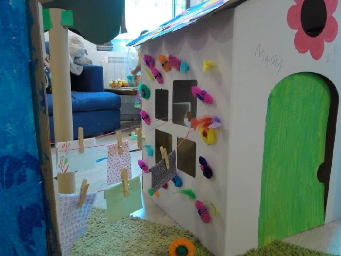 A Mondoko cardboard playhouse with a green door, decorated by children with paper flowers and pegs.